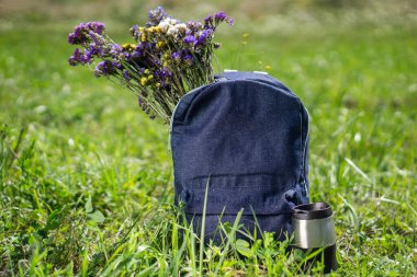 Rahatlama ve yaz yürüyüşü konsepti. Mavi kot pantolon okul çantası yeşil çimlerin üzerinde duruyor. Yanında seyahat kupası ve içinde güzel çayır çiçekleri var.