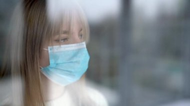 Koruyucu tıbbi maskeli genç, çekici bir sarışın pencereye bakıyor ve elleriyle pencereye dokunuyor. Coronavirus covid-19 izolasyon kavramı.