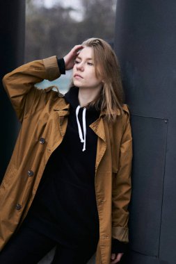 Uzun saçlı, turuncu pelerinli ve siyah kapüşonlu çıplak makyajlı çekici genç kız portresi karanlık şehir arka planına sahip. Yaşam tarzı ve moda konsepti.