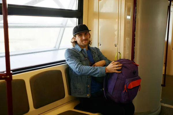 Happy young bearded traveler man in baseball hat and jeans jacket with purple backpack sitting inside metro train in european country. Tourism, traveling, underground or lifestyle concept.