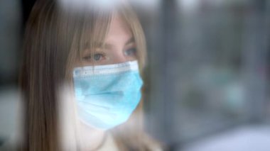 Güzel sarışın ev izolasyonu sırasında koruyucu sağlık maskesi takmış camdan dışarı bakıyor ve elleriyle pencereye dokunuyor. Coronavirus konsepti.