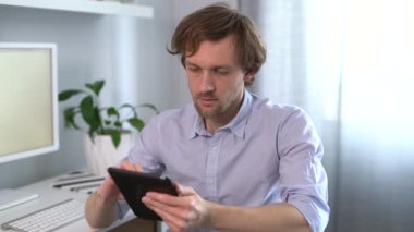 Kapalı sakallı erkek, tablet kullanıyor. İPad rahatlatıcı ve gülümseyen bir sandalyeye oturup bilgisayar ve parlak bir altyapıya sahip olmak. Mesajlaşma, internette gezinme, online alışveriş veya karantina konsepti