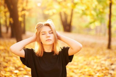 Siyah tişörtlü genç sarışın kız sonbahar parkında elleri ve gözleri kapalı poz veriyor. Gençlik portresi, model görünüm kitabı ya da sonbahar sonbahar konsepti