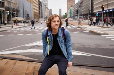 Siyah tişörtlü ve kot ceketli sıradan bir genç metal çitin üzerinde oturuyor ve büyük şehir arka planına sahip kameraya bakıyor. Seyahat ve yaşam tarzı kavramı.