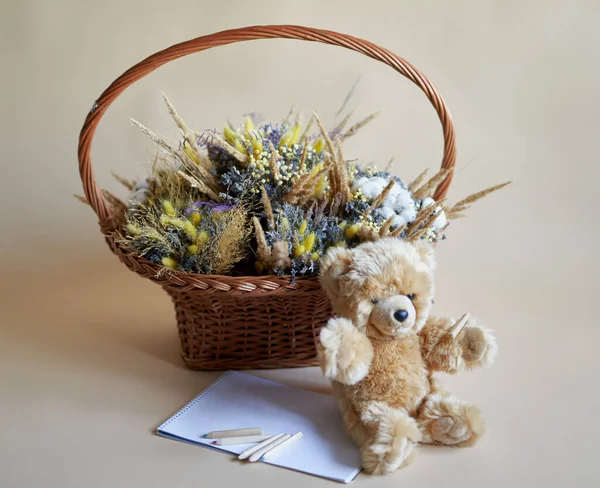 柳条篮里有一束干花 旁边有泰迪熊 还有笔记本和铅笔 照片上的米色背景 — 图库照片