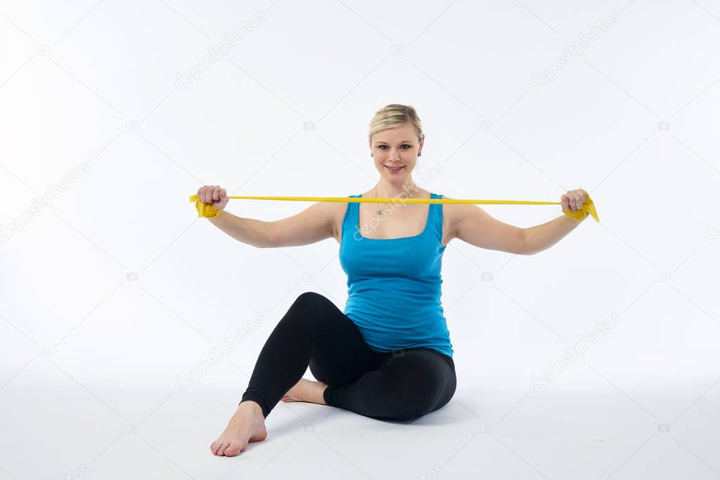 Young blondy pregnant woman in blue tank top does exercises with yellow fitness rubber band, white background