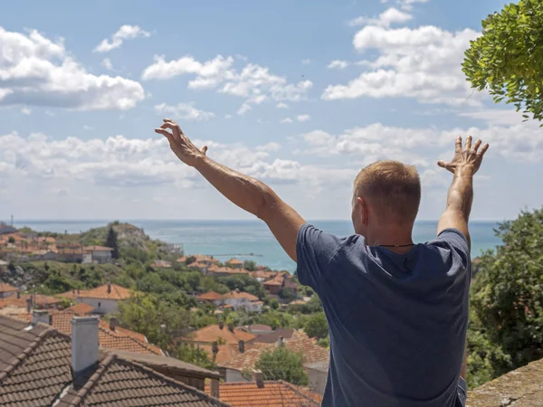 Un hombre con los brazos extendidos sobre una ciudad con techos de baldosas mira al mar . — Foto de Stock