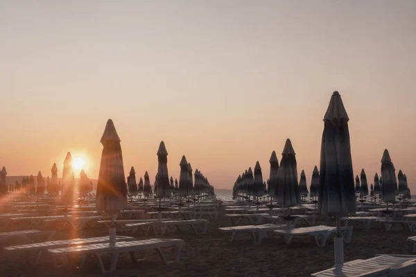 Sonnenaufgang am Strand. Liegestühle und Sonnenschirme in den Strahlen der aufgehenden Sonne. Strand ohne Menschen. — Stockfoto
