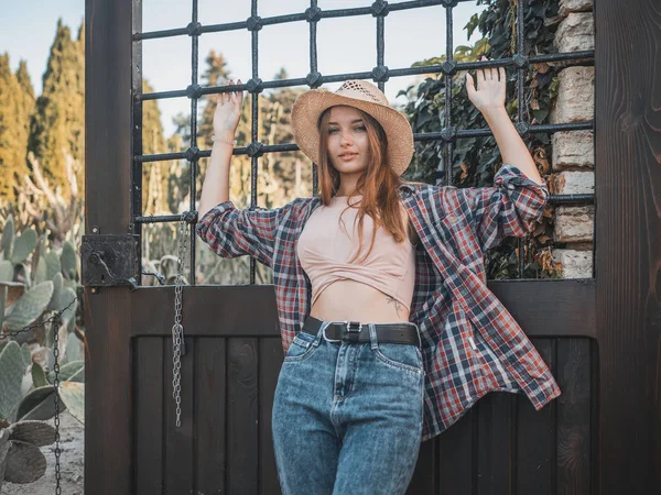 Joven mujer pelirroja en un rancho con un sombrero de paja, una camisa a cuadros y jeans en el fondo de la parrilla. Estilo casual —  Fotos de Stock