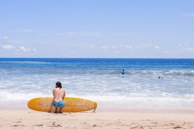 Kum plajındaki okyanusa bakarak otururken sörfçü