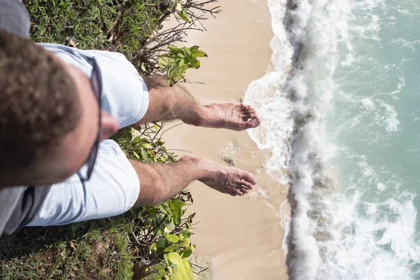 Un hombre colgó sus piernas en el borde de un acantilado se sienta sobre el océano . — Foto de Stock