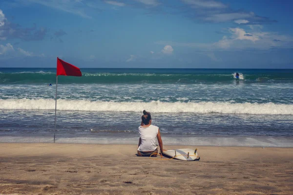 Una joven con una tabla de surf se sienta en la playa y mira las olas — Foto de Stock