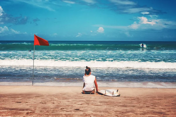 Una joven con una tabla de surf se sienta en la playa y mira las olas — Foto de Stock