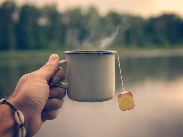 Männliche Hand mit einem Becher Tee auf dem Hintergrund des Sees — Stockfoto