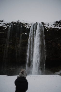 Bir kişi bir şapka kullanılan bir eşarp ile kışın ortasında Seljalandsfoss, İzlanda önünde duruyor.