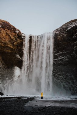 Buz Skogafoss, İzlanda önünde bir yama üzerinde sarı ceket ayakta olan bir kişi.