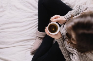 Yünlü kazaklı bir kadın yatakta oturmuş, kahve içiyor. Resim yukarıdan çekilmiş.