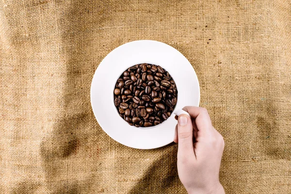 A hand reaching for a mug filed with coffee beans on a brown, structured background