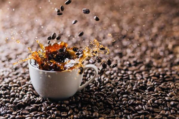 コーヒー豆が完全にコーヒー豆で構成されている背景にコーヒーで満たされたマグカップに飛び散るコーヒー豆 — ストック写真