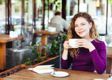 Güzel çekici genç sarışın kadın kapuçino içiyor ve kruvasan şehir kafede yaz gün yemek kıvırcık saçlı. Defter ve telefon masaya vardır