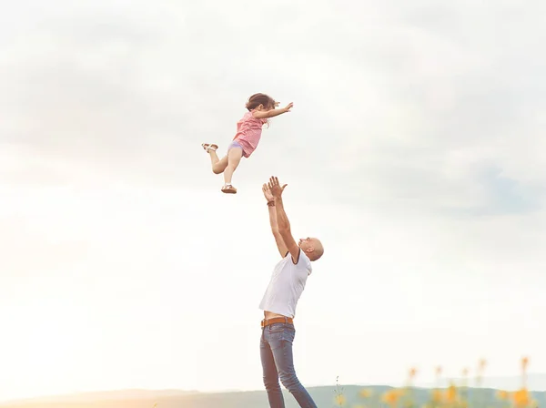 Mutlu baba kızını güneşli alan ile günbatımı üzerinde oynarken. Yaz mutlu aile yaşam tarzı, gerçek duygular. Küçük kız yüksekten uçuyor gökyüzünde babamın elinden