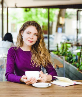 Güzel çekici genç sarışın kadın kapuçino içiyor ve kruvasan şehir kafede yaz gün yemek kıvırcık saçlı. Defter ve telefon masaya vardır