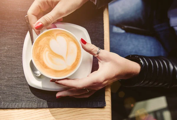 Frauenhände mit roter Maniküre halten Tasse Cappuccino auf Tisch im Café. — Stockfoto