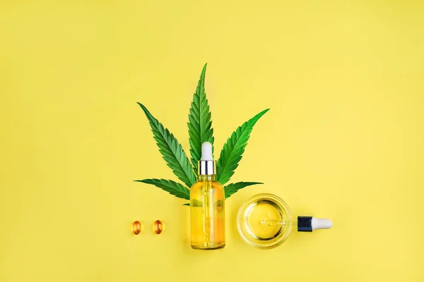 Flaska med CBD-olja, pipett, kapslar och cannabisblad på gul bakgrund. Minimalism, Flat Lay. — Stockfoto