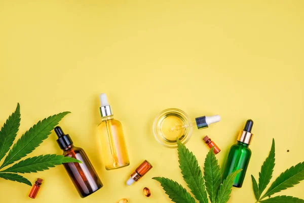 Verschillende glazen flessen met CBD olie, THC tinctuur en cannabis bladeren op gele achtergrond. Platte lay, minimalisme. Cosmetica CBD olie. — Stockfoto