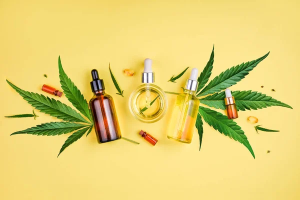 Different glass bottles with CBD OIL, THC tincture and cannabis leaves on yellow background. Flat lay, minimalism. Cosmetics CBD oil. — Stock Photo, Image