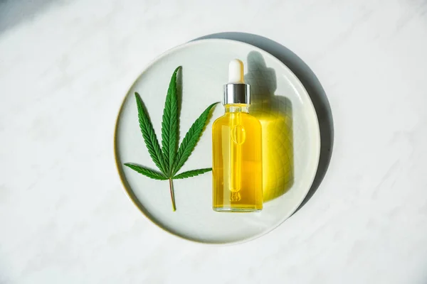 CBD olje flaska och pipett på en liten tallrik och på en marmor yta. Cannabis lämnar i närheten. — Stockfoto