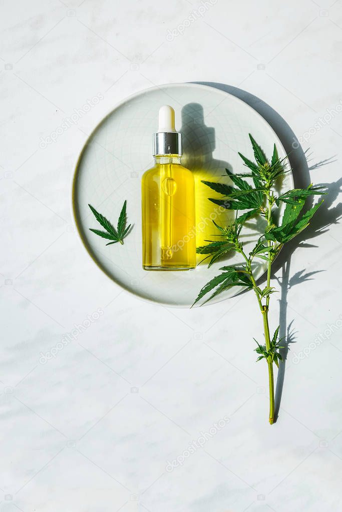 CBD Oil bottle and dropper on a small plate and on a marble surface. Cannabis leaves nearby.