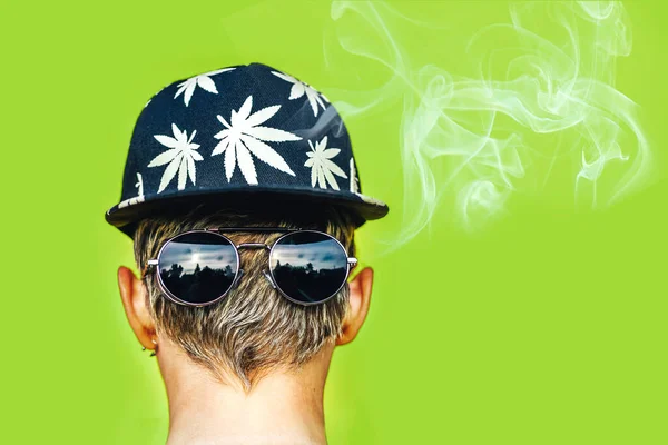 Meisje met zonnebril en pet met bladeren van marihuana rookt op groene achtergrond — Stockfoto