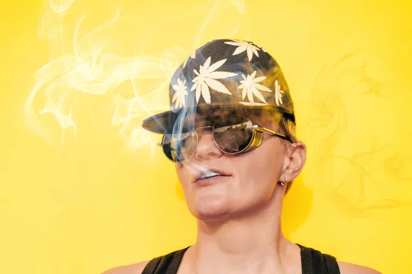 Dziewczyna w okularach przeciwsłonecznych i czapce z liści marihuany pali na żółtym tle — Zdjęcie stockowe