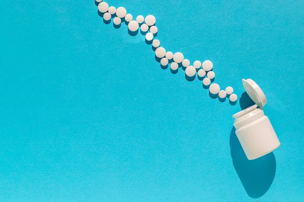 Beyaz haplar mavi arka plandaki şişeden düşüyor. Tıbbi düzlük