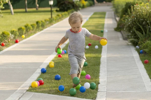 Милый мальчик играет с красочными мячами в зеленой траве в саду, парке или на заднем дворе — стоковое фото