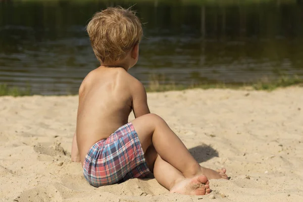 Dwuletnie dziecko chłopiec siedzi na piasku i plażowanie, patrząc od hotelu, widok z tyłu. Koncepcję wakacji — Zdjęcie stockowe