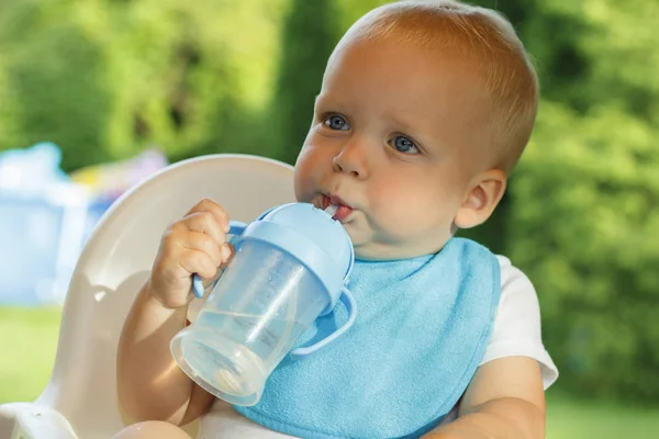 Забавный малыш пьет воду из бутылки. Он стоит на зеленой лужайке. — стоковое фото