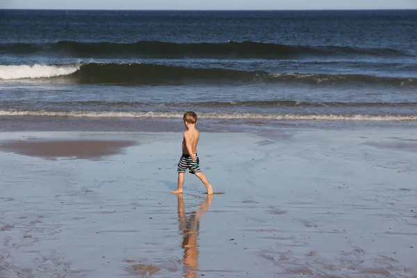 Ένα μικρό ξανθό αγοράκι περπατά κατά μήκος της ακτής του ωκεανού μόνο χωρίς γονείς. Καλοκαίρι, ζεστό, θάλασσα, ήλιος, παραλία. — Φωτογραφία Αρχείου