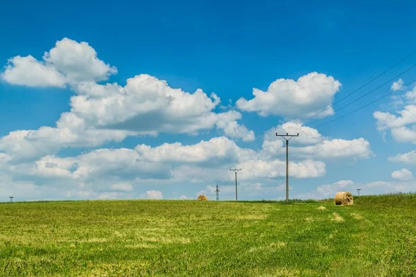 Rural countryside with harvested agriculture field, straw bales, electricity poles and lines on horizon, sunny summer day, bright blue sky with white clouds