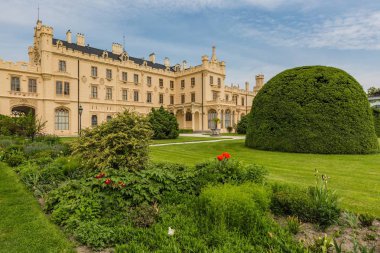 Lednice, Çek Cumhuriyeti - 27 Mayıs 2019: Güney Moravya'da sarı cepheli ünlü Lednice kalesi. Ön planda yeşil çim, çalı ve çiçek ile Bahçe. Güneşli bahar günü, mavi gökyüzü, beyaz bulutlar.