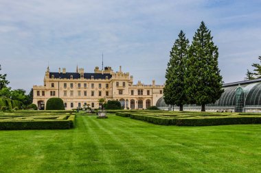 Lednice, Çek Cumhuriyeti - 27 Mayıs 2019: Güney Moravya'da sarı cepheli ünlü Lednice kalesi. Yeşil çimler, çalılar, ağaçlar ve sera ile Bahçe. Güneşli bahar günü, mavi gökyüzü, beyaz bulutlar.