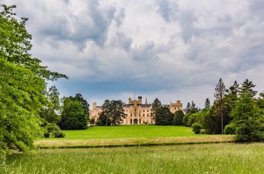 Lednice, Çek Cumhuriyeti - 27 Mayıs 2019: Güney Moravya'daki Lednice kalesinin görünümü, yeşil çayır üzerinde sarı cepheli ve yan tarafta ağaçlar. Güneşli bahar günü, beyaz bulutlarla dramatik mavi gökyüzü.
