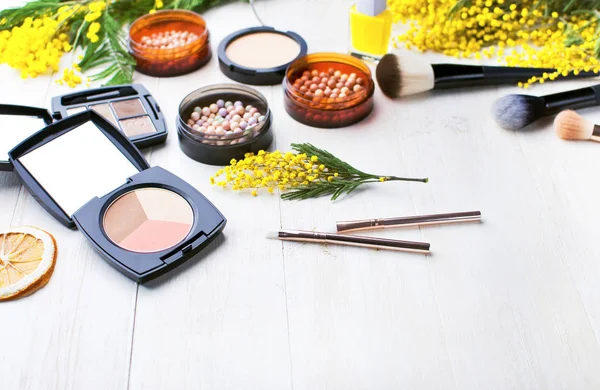 Set of decorative cosmetics for make-up Powder Rouge Eyeshadow Corrector Brushes and flowers of mimosa on a light wooden background. Makeup Accessories with copy space.