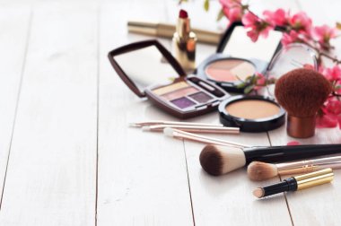 Kopya alanı ile beyaz ahşap zemin üzerinde pembe çiçekli makyaj için çeşitli kozmetik ürünleri. Kalıcı makyaj, ruj, rimel, göz farı, toz, fırçalar