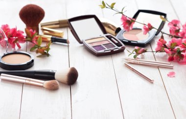 Kopya alanı ile beyaz ahşap zemin üzerinde pembe çiçekli makyaj için çeşitli kozmetik ürünleri. Kalıcı makyaj, ruj, rimel, göz farı, toz, fırçalar