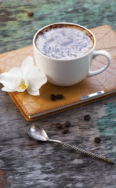 A cup of hot coffee and an orchid flower with a diary on a wooden background. Traditional drink of cappuccino or cocoa Wooden background Orchid Notepad Diary Autumn drink
