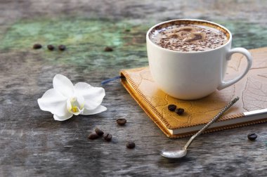 Bir fincan sıcak kahve ve ahşap arka plan üzerinde bir günlük ile bir orkide çiçeği. Cappuccino veya kakao ahşap arka plan orkide Not defteri günlüğü sonbahar içki geleneksel içki
