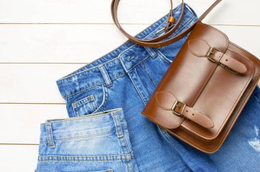 Üstten görünüm kot pantolon, kahverengi deri çanta ve hafif ahşap arka plan üzerinde bir deri kemer. Güzel kıyafetleri ve aksesuarları ayrıntılarını. Kot doku veya kot arka plan. Giyim, moda