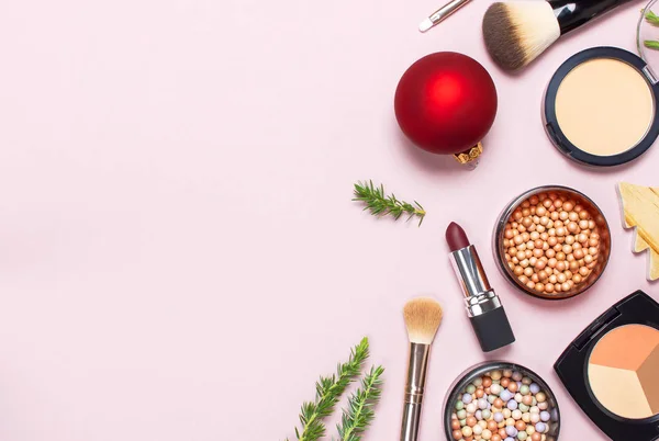 Various cosmetic products for make-up, blusher, powder, brushes, lipstick, eyeshadow and Christmas gift, fir branches, red ball on pink background Flat lay top view New year concept winter decoration